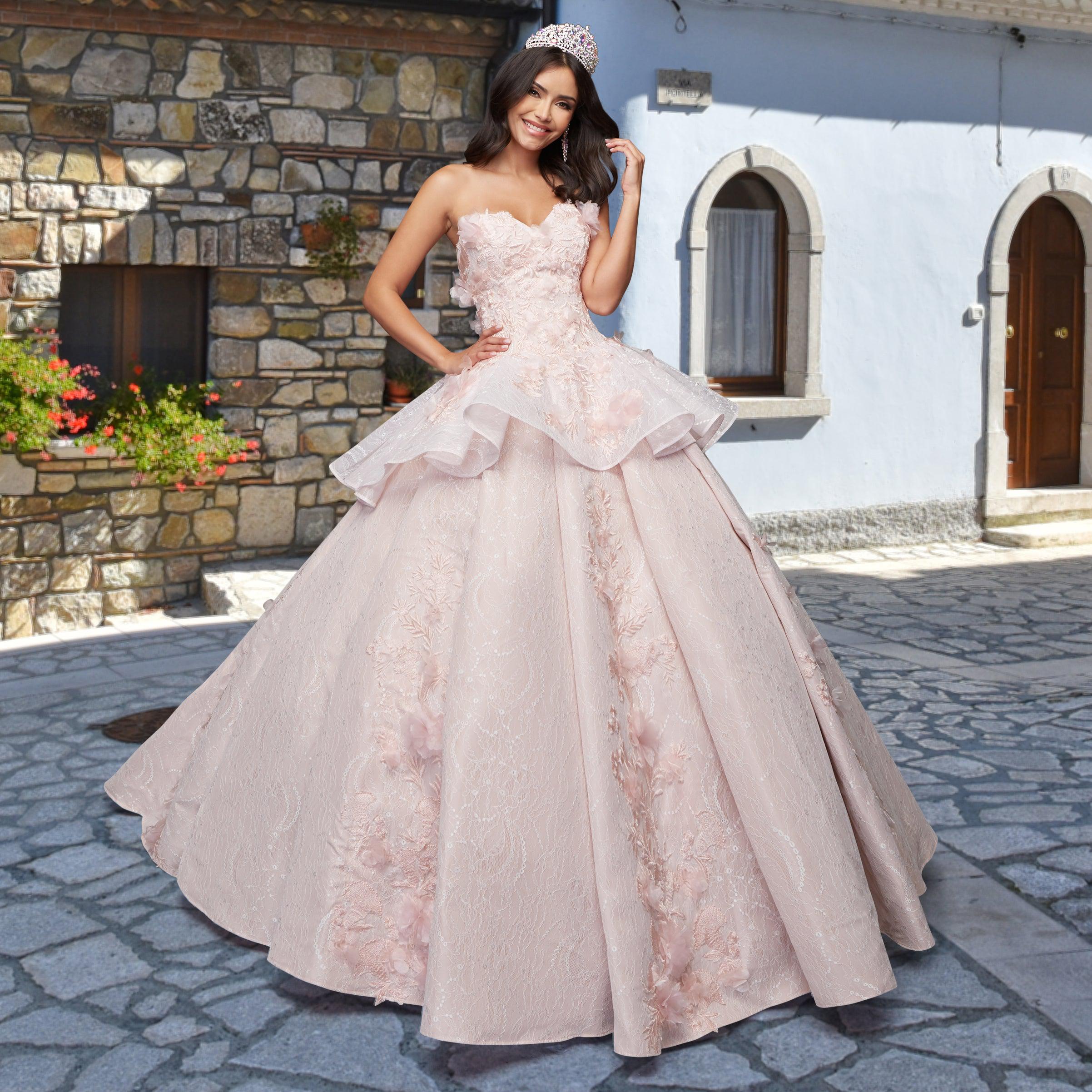 A woman in the Quinceañera Sample Dress QF115 and a tiara smiles on a cobblestone street, standing before a stone and stucco building.